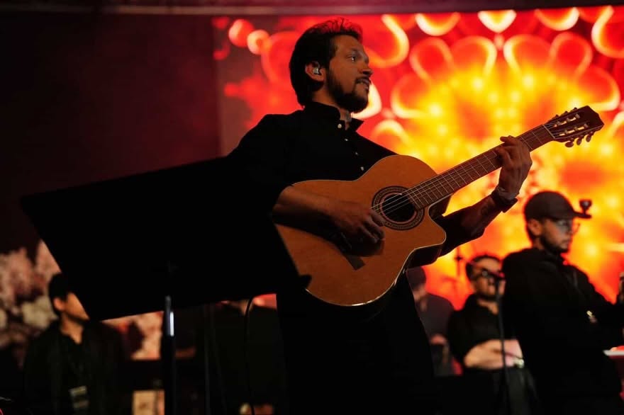 Glen Fernandez playing acoustic guitar in front of a red backdrop